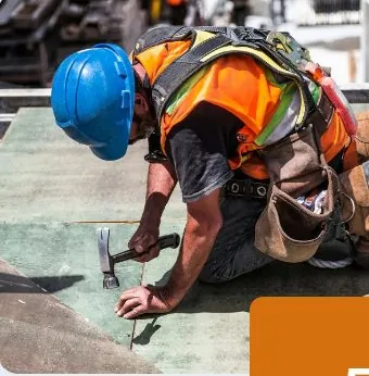 Roofer in a blue hard hat and orange safety vest hammering on a roof